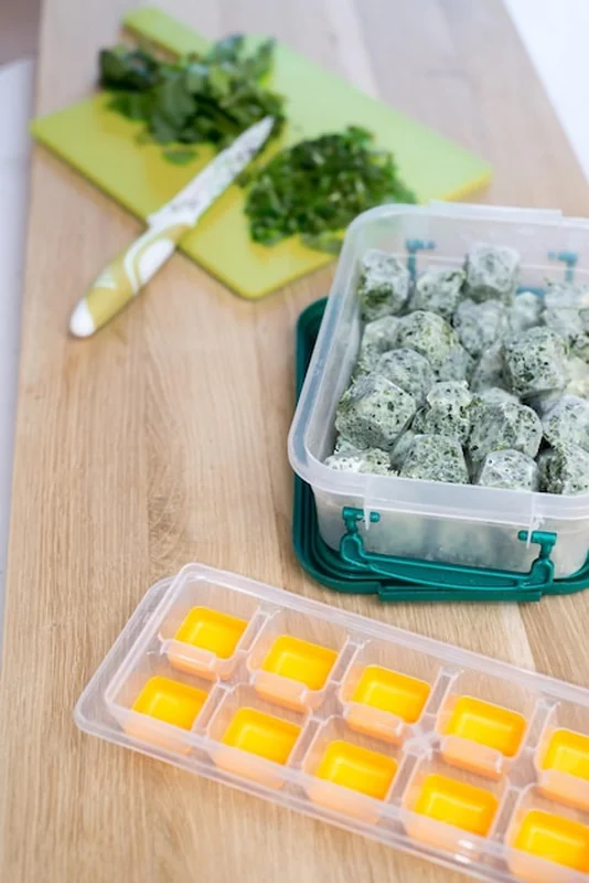 Mrożone warzywa, deska do krojenia i pojemnik z mrożonymi kostkami ziół w oliwie z oliwek. Widok z góry